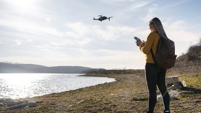 A woman wearing a yellow sweater and backpack operates a small drone with a remote controller near a lakeside, with hills and a partly cloudy sky in the background.