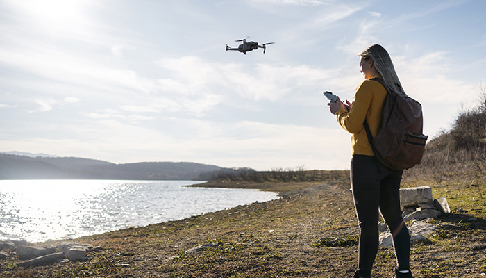 A woman wearing a yellow sweater and backpack operates a small drone with a remote controller near a lakeside, with hills and a partly cloudy sky in the background.