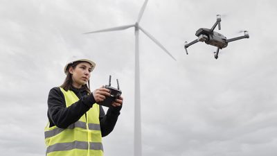 A female engineer wearing a high-visibility safety vest and hard hat operates a drone using a remote controller. A wind turbine is visible in the background under a cloudy sky, symbolizing the use of drone technology in renewable energy inspections.