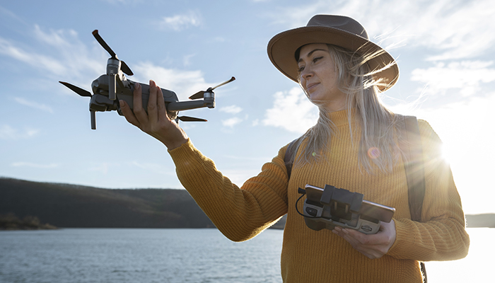 A woman wearing a brown hat and mustard yellow sweater holds a drone in one hand and a remote controller in the other, standing near a body of water with hills in the background under a clear blue sky.