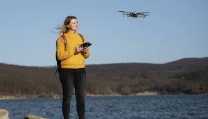 A young woman in a mustard yellow sweater and black jeans is flying a small drone near a lake with hills in the background on a clear day.