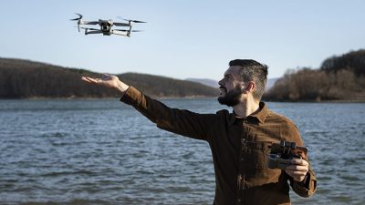 A man operating a drone near a lake, extending his hand as the drone hovers in front of him, with forested hills in the background under a clear blue sky.