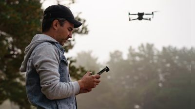 A man wearing a denim jacket and black cap is controlling a drone with a remote controller outdoors, surrounded by trees and misty weather.