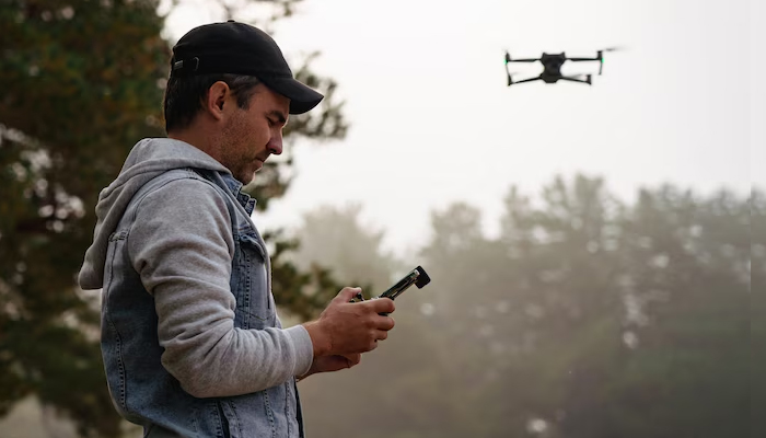 A man wearing a denim jacket and black cap is controlling a drone with a remote controller outdoors, surrounded by trees and misty weather.