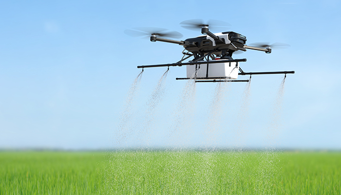An agricultural drone equipped with multiple rotors flying over a green field, spraying a fine mist of liquid evenly across the crops under a clear blue sky.