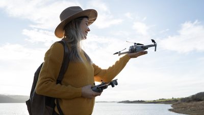 A woman wearing a brown hat and mustard yellow sweater is holding a remote control in one hand and a small drone in the other. She stands outdoors near a body of water with a scenic background of hills under a partly cloudy sky.
