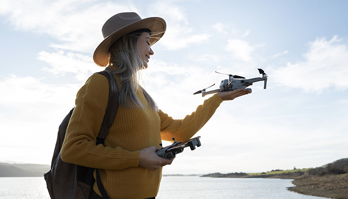 A woman wearing a brown hat and mustard yellow sweater is holding a remote control in one hand and a small drone in the other. She stands outdoors near a body of water with a scenic background of hills under a partly cloudy sky.