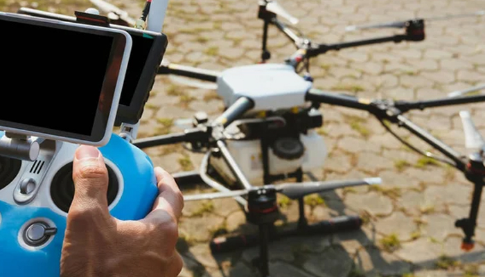 Close-up of a person holding a blue drone remote controller with two display screens, preparing to operate a large multi-rotor drone resting on the ground with a mix of grass and paved surface.