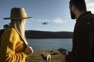 A man and woman outdoors by a lake operating a drone; the woman is wearing a wide-brimmed hat and a yellow sweater, while the man is holding a drone controller. A dog is standing on the grass near them, and the drone is flying above the water with hills in the background under a clear sky.