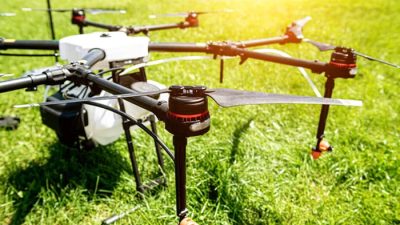 A close-up view of a large agricultural drone with multiple propellers standing on green grass, illuminated by sunlight, highlighting its mechanical components and rotor blades.