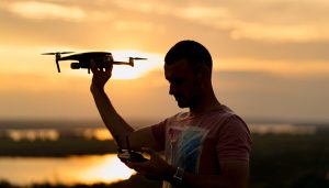 A man holding a drone in one hand and a remote controller in the other, silhouetted against a scenic sunset with a river and landscape in the background.