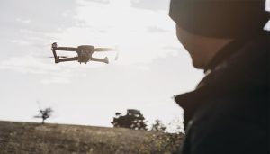 A person in a dark jacket and beanie watches a drone flying in an open field during daylight. The sun is partially behind the drone, casting a silhouette effect, with trees and dry grass in the background.