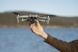 A person holding a compact, gray surveillance drone with a camera, poised for takeoff over a body of water with a blurred natural landscape in the background.