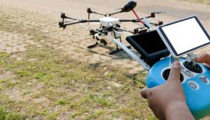Close-up of a person holding a blue drone remote controller with two display screens, preparing to operate a large multi-rotor drone resting on the ground with a mix of grass and paved surface.