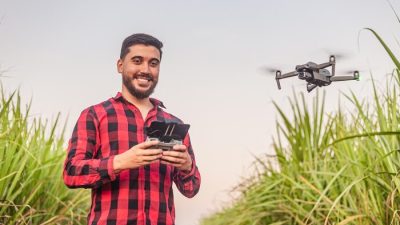 A smiling man in a red and black checkered shirt operates a drone using a remote controller while standing in a green sugarcane field, demonstrating agricultural drone use for farming.