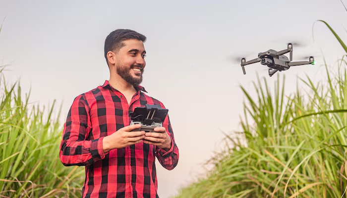 A young man in a red checkered shirt smiles while operating a drone with a remote control in the middle of a lush green agricultural field, showcasing the benefits of drone technology in farming.