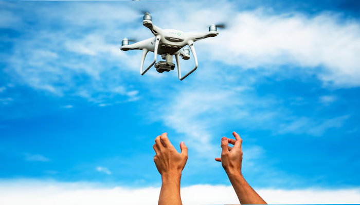 A white quadcopter drone hovers in the sky above a pair of outstretched hands, set against a bright blue sky with scattered clouds.