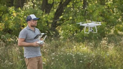 A man wearing a cap and sunglasses operates a white quadcopter drone using a remote controller with a screen, standing in a grassy field with trees in the background.