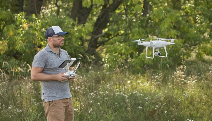 A man wearing a cap and sunglasses operates a white quadcopter drone using a remote controller with a screen, standing in a grassy field with trees in the background.